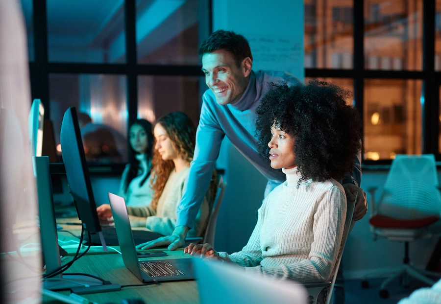 Vrouw met kroes haar overlegt met collega achter computer op een modern kantoor in de avond.